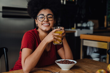 Smiling african woman drinking orange juice and eating cereal while sitting in kitchen