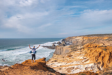 Joyful expression of a young backpacker standing on the edge of a cliff overlooking the Atlantic Ocean in the Odemira region, western Portugal. Wandering along the Fisherman Trail, Rota Vicentina