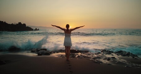 Fototapeta na wymiar Woman silhouette raises hands up on black sand beach at sunset. Twilight orange sky. Adult girl relax watching powerful sea waves landscape.