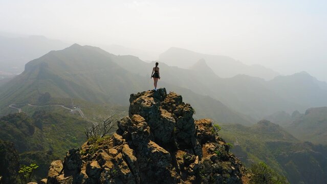 Aerial shooting from drone of a woman tourist is enjoying scenery of green mountains. Young hiking girl stands on top of the cliff. Time to trip. Go everywhere. Strength of women. Girls power.