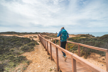 Fototapeta na wymiar Young and courageous Vagabund roaming the Portuguese countryside. A backpacker walks along a wooden walkway in the Odemira region of Portugal. Fisherman Trail, Rota Vicentina