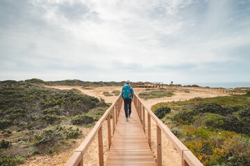 Fototapeta na wymiar Young and courageous Vagabund roaming the Portuguese countryside. A backpacker walks along a wooden walkway in the Odemira region of Portugal. Fisherman Trail, Rota Vicentina