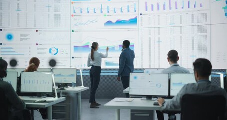 Black Male Financial Data Analysts And Caucasian Female Project Manager Having A Meeting In Monitoring Office With Analytics Feed On Big Digital Screen. Consulting Firm With Employees Behind Computers