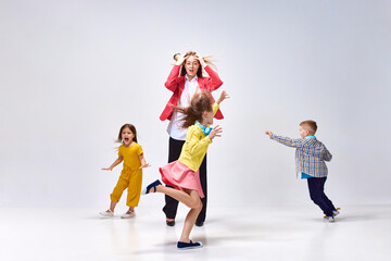 Portrait of tired young woman, mother holding head with hands around little noisy kids playing against grey studio background. Concept of family, motherhood, childhood, fashion, lifestyle