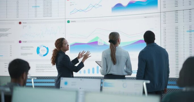 Diverse Team Of Data Scientists Having A Meeting And Discussing Reports In a Modern Monitoring Office With Analytics Feed On a Big Digital Screen. Deep Learning Company With Employees Behind Computers
