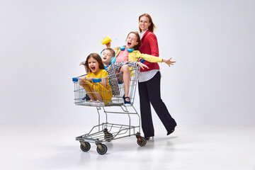 Portrait of happy young woman, mother going shopping with her little kids, children sitting in shopping trolley against grey studio background. Concept of family, motherhood, childhood, lifestyle