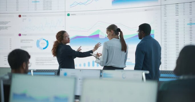 Diverse Team Of Data Scientists Having A Meeting And Discussing Reports In Monitoring Office With Analytics Feed On Big Digital Screen. Artificial Intelligence Company With Employees Behind Computers