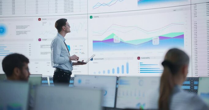 Male Senior Data Analyst Reviewing Financial Information And Reports on Big Digital Screen in Monitoring Office. Diverse Employees Working Behind Desktop Computers In Venture Capital Company.