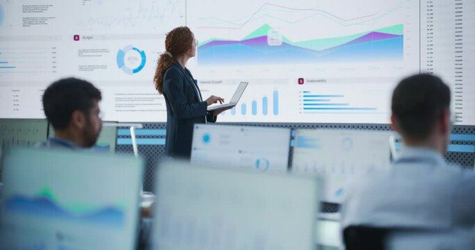 Female Project Manager Using Laptop And Analyzing Information Graphs On Big Digital Screen In Monitoring Office. Hispanic Woman Working For Big Data Company, Diverse Employees Behind Desktop Computers