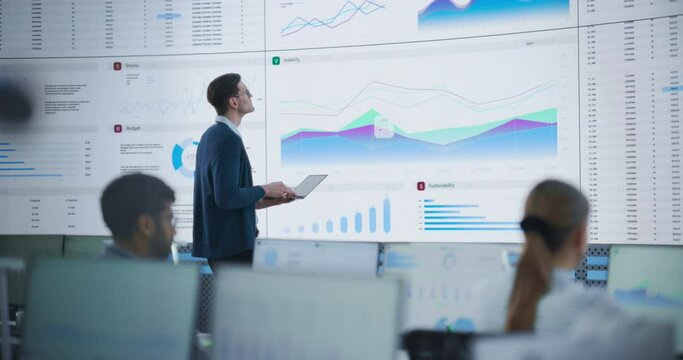 Male Senior Financial Analyst Reviewing Information And Reports on Big Digital Screen in Modern Monitoring Office. Diverse Employees Working Behind Desktop Computers In Investment Management Company.