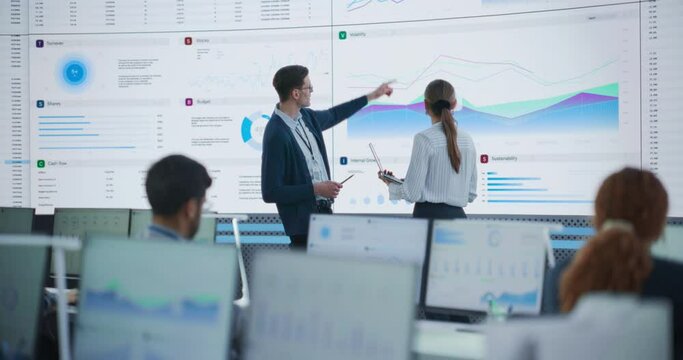 Male Data Scientist And Female Project Manager Discussing Reports And Graphs On Big Digital Screen In Modern Monitoring Office. Diverse Employees Working Behind Computers in Deep Learning Company.