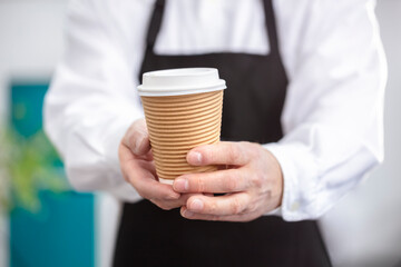 Waiter barista holding a take out paper disposable cup of hot coffee in cafe