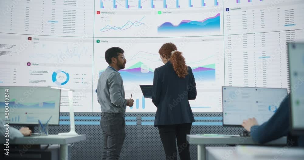 Poster Back View Of Indian Data Analyst And Hispanic Manager Discussing Reports On Digital Screen in Monitoring Room. Male and Female Experts Talking, Employees Working On Computers In Consulting Company