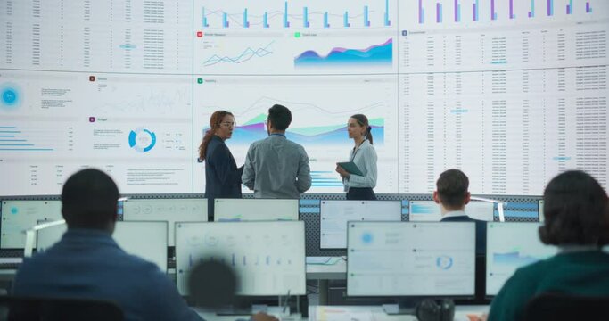 Wide Back Shot of Diverse Team of Data Scientists Discussing Graphs And Reports On Big Digital Screen In Monitoring Office. Multiethnic Employees Working On Desktop Computers For Deep Learning Company