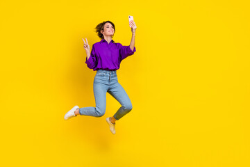 Fototapeta na wymiar Full length photo of excited pretty woman dressed purple shirt jumping recording video vlog v-sign empty space isolated yellow color background
