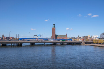 Fototapeta na wymiar Blue commuter train on a bridge over the river Strömmen in front of the Town City Hall, a sunny spring day in Stockholm