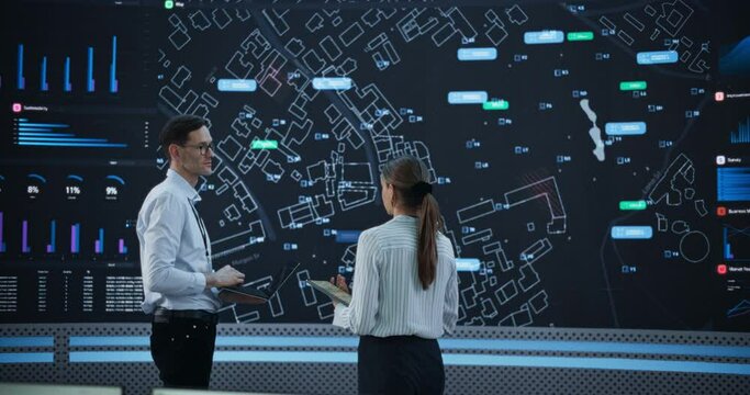Male And Female Data Analysts Discussing New Locations For Business In Front Of Big Digital Screen With City Map In Monitoring Office. Diverse Employees Working Behind Computers For Consulting Company