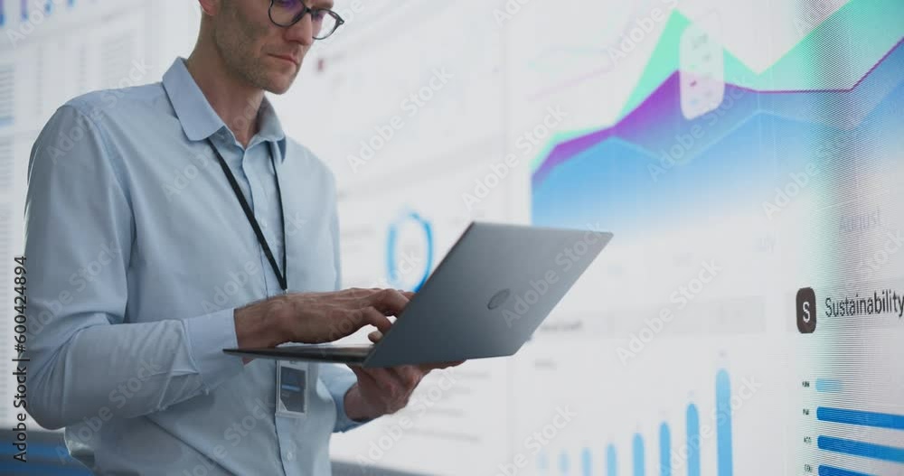 Poster Close Up Of Successful Caucasian Man Typing On Laptop Computer And Standing Next To Big Digital Wall With Data In The Office. Male Business Analyst Creating A Growth Strategy For Innovative Startup.