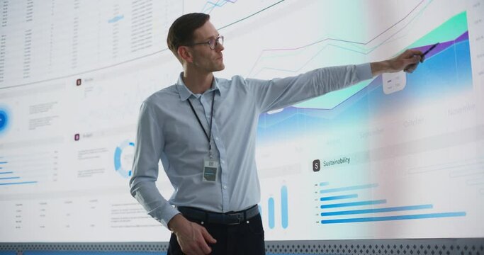 Male CEO Of Data Science Company Is Giving Presentation In Front Of Big Digital Screen With Graphs And Charts. Successful Caucasian Man Reviewing Business Accomplishments And Objectives In the Office.