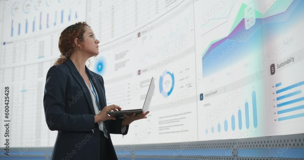 Poster In the Monitoring Office: Successful Hispanic Businesswoman Holding Laptop Computer And Analyzing Data On Big Digital Screen With Graphs and Charts. Female IT Entrepreneur Running Software Company.