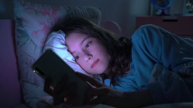 Teen girl lying in bed at night using smartphone, scrolling social media and texting. Not healthy sleep. Insomnia and the harm of the smartphone before going to bed. Dependence on social networks