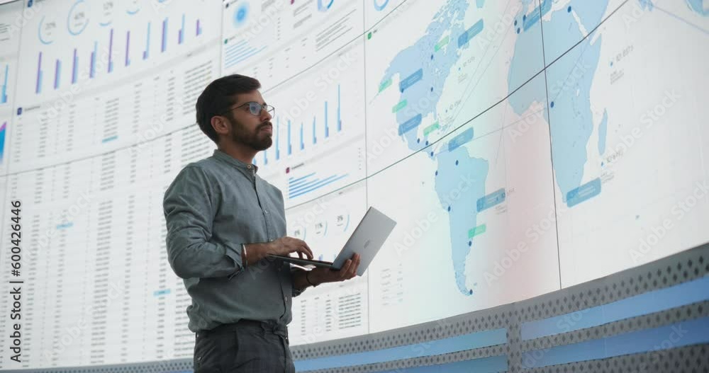 Poster indian male product distribution expert holding laptop computer, analyzing world map on big digital 