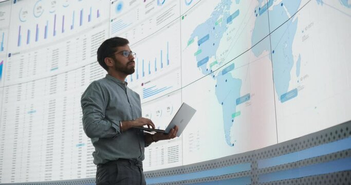 Indian Male Product Distribution Expert Holding Laptop Computer, Analyzing World Map On Big Digital Screen In Monitoring Office. Successful Man Looking For Efficient Routes For International Delivery.