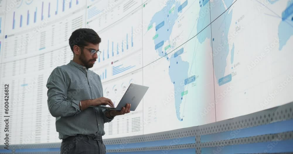 Leinwandbilder indian male logistics expert holding laptop computer and analyzing world map on big digital screen i