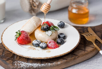 Cottage cheese pancakes, cheesecakes with fresh blueberries, strawberries, honey and mint leaves on a stylish wooden board on a gray concrete table. Natural products. Healthy and delicious breakfast.
