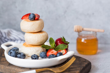 Cottage cheese pancakes, Russian cheesecakes with fresh blueberries, strawberries, honey and mint leaves on a stylish wooden board on a gray concrete table.  Healthy and delicious breakfast.