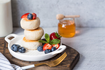 Cottage cheese pancakes, Russian cheesecakes with fresh blueberries, strawberries, honey and mint leaves on a stylish wooden board on a gray concrete table.  Healthy and delicious breakfast.