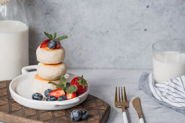 Cottage cheese pancakes, Russian cheesecakes with fresh blueberries, strawberries and mint on a stylish wooden board on a gray concrete table. Natural products. Healthy and delicious breakfast.