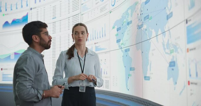 Caucasian Female Regional Manager Talking To Indian Male Distribution Expert In Front Of Big Digital Screen With Infographics And Map. Successful Man And Woman Discussing New Markets For Expansion.