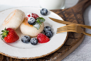 Cottage cheese pancakes, Russian cheesecakes with fresh blueberries, strawberries and mint on a stylish wooden board on a gray concrete table. Natural products. Healthy and delicious breakfast.