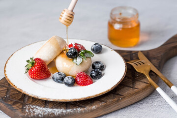 Cottage cheese pancakes,cheesecakes with fresh blueberries, strawberries, honey and mint leaves on a stylish wooden board on a gray concrete table. Natural products. Healthy and delicious breakfast.