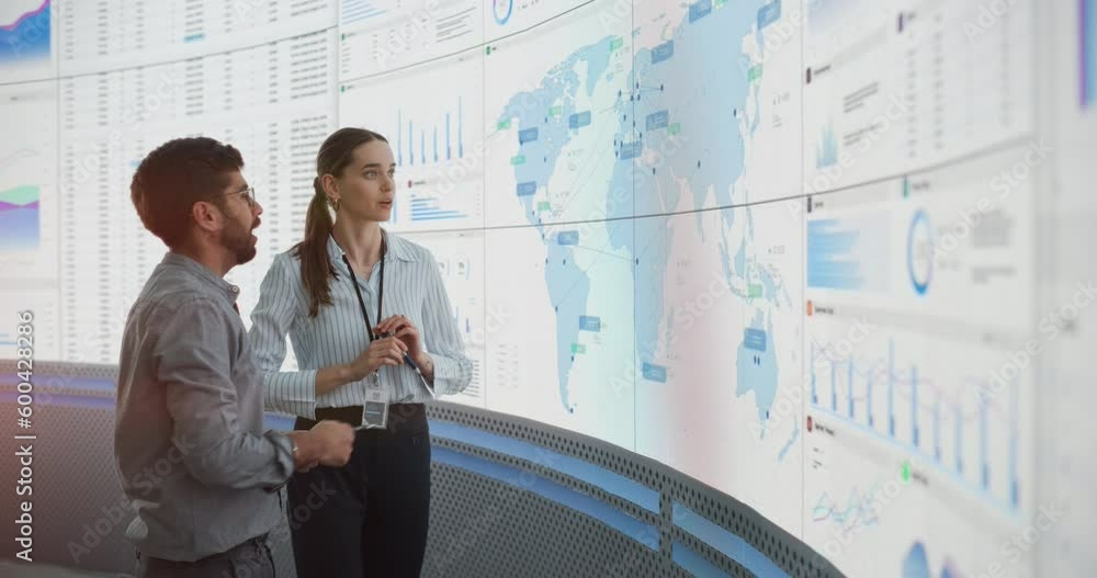 Poster caucasian female manager talks to indian male distribution expert in front of big digital screen wit