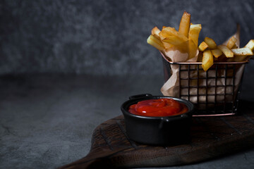 French fries in a metal basket with salt and ketchup on a stylish board on a dark background. Fried...