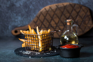 French fries in a metal basket with salt, ketchup and sunflower oil on a stylish board on a dark...