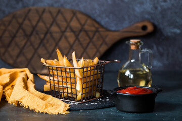 French fries in a metal basket with salt, ketchup and sunflower oil on a stylish board on a dark...