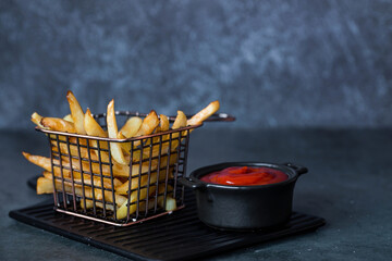 French fries in a metal basket with salt and ketchup on a stylish board on a dark background. Fried...