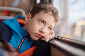 a cute boy in a bright sport jacket is riding alone on a tram and looks out the window. The boy's face is reflected in the window. Close-up
