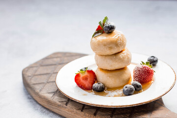 Cottage cheese pancakes, cheesecakes with fresh blueberries, strawberries, honey and mint leaves on a stylish wooden board on a gray concrete table. Natural products. Healthy and delicious breakfast.