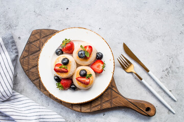 Cottage cheese pancakes, cheesecakes with fresh blueberries, strawberries, honey and mint leaves on a stylish wooden board on a gray concrete table. Natural products. Healthy and delicious breakfast.