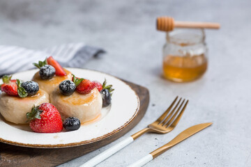 Cottage cheese pancakes, cheesecakes with fresh blueberries, strawberries, honey and mint leaves on a stylish wooden board on a gray concrete table. Natural products. Healthy and delicious breakfast.