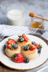 Cottage cheese pancakes, cheesecakes with fresh blueberries, strawberries, honey and mint leaves on a stylish wooden board on a gray concrete table. Natural products. Healthy and delicious breakfast.