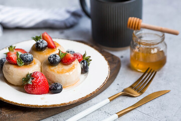 Cottage cheese pancakes, cheesecakes with fresh blueberries, strawberries, honey and mint leaves on a stylish wooden board on a gray concrete table. Natural products. Healthy and delicious breakfast.