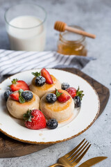 Cottage cheese pancakes, cheesecakes with fresh blueberries, strawberries, honey and mint leaves on a stylish wooden board on a gray concrete table. Natural products. Healthy and delicious breakfast.