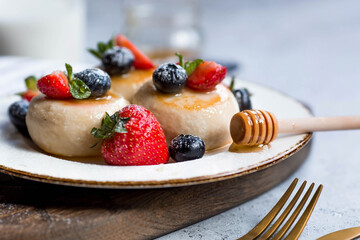 Cottage cheese pancakes, cheesecakes with fresh blueberries, strawberries, honey and mint leaves on a stylish wooden board on a gray concrete table. Natural products. Healthy and delicious breakfast.