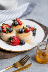 Cottage cheese pancakes, cheesecakes with fresh blueberries, strawberries, honey and mint leaves on a stylish wooden board on a gray concrete table. Natural products. Healthy and delicious breakfast.