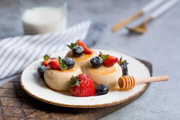 Cottage cheese pancakes, cheesecakes with fresh blueberries, strawberries, honey and mint leaves on a stylish wooden board on a gray concrete table. Natural products. Healthy and delicious breakfast.